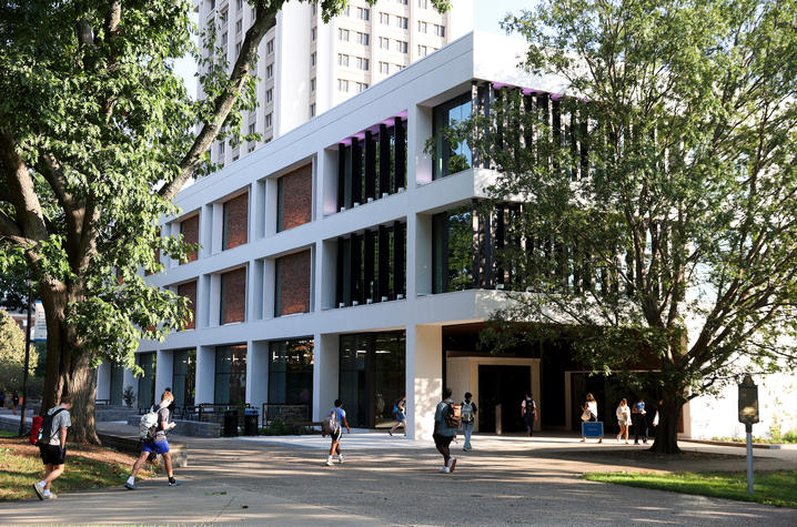 Students walk along a shaded campus pathway outside a modern academic building surrounded by large trees.