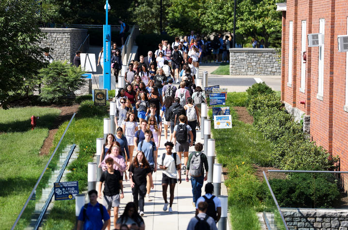 A large group of students walks along a campus sidewalk between academic buildings on a sunny day.