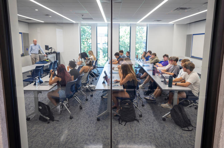 Students sit at tables using laptops while an instructor leads a class in a modern classroom with large windows and bright lighting.