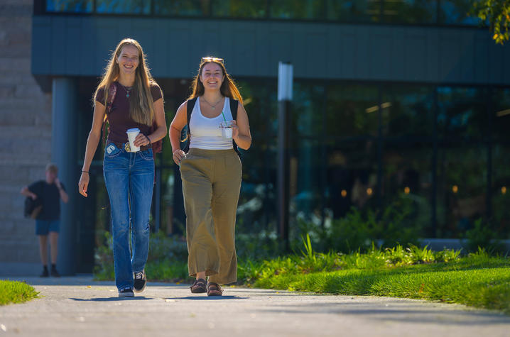 photo of students walking Two students walk along a campus sidewalk carrying backpacks and drinks, with a modern academic building and landscaped greenery in the background.