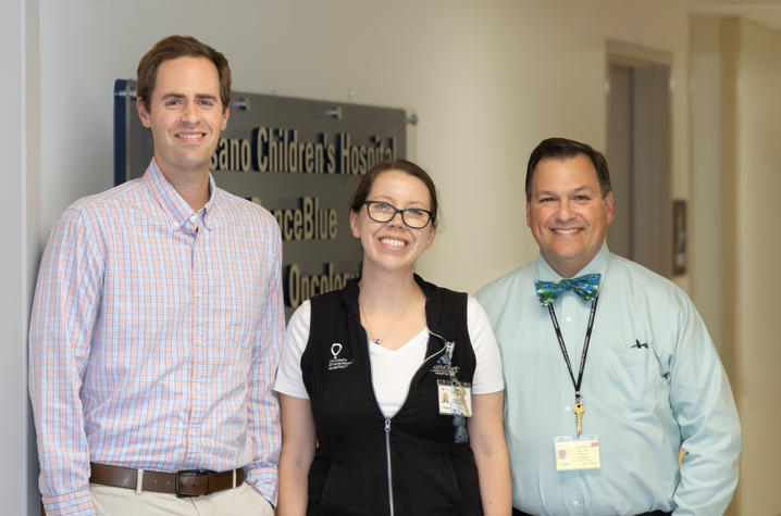 James Collard, Amanda Harrington, M.D., and John A. D’Orazio, M.D., Ph.D. Three healthcare team members stand together in a hospital hallway in front of a sign for a pediatric oncology clinic. One person wears a plaid button-down shirt, another wears a black hospital vest with an ID badge, and the third wears a light blue shirt