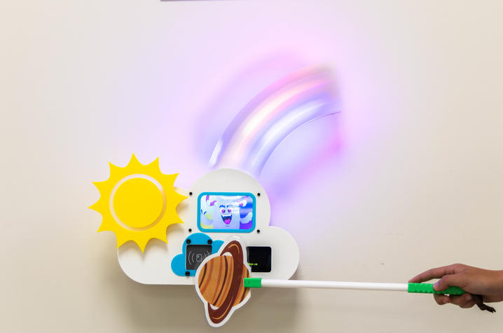 Interactive station for walking test A hand holds a planet-themed wand in front of a colorful wall-mounted game station featuring a lighted rainbow and animated character.