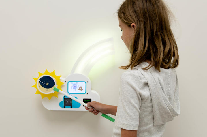 Child demonstrates gamified walking test A child uses a moon-themed wand at a lighted wall-mounted game station displaying an animated character and the number four.