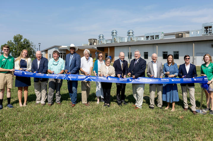 Ribbon Cutting A group of people standing outdoors in front of a large modern industrial or educational facility, holding a blue ceremonial ribbon and scissors during a ribbon-cutting event. The building features metal siding and rooftop ventilation equipment.
