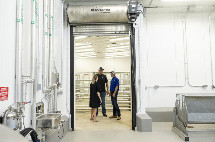 MFIAC processing space Interior view of a livestock handling and processing facility with white walls, exposed utility pipes and bright overhead lighting. Three people stand and talk in a doorway beneath a large stainless steel roll-up garage door labeled “Raynor Garage Doors.”