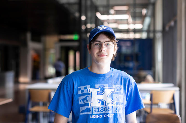 A person wearing a University of Kentucky cap and blue K Week t-shirt stands in a campus study area with tables and chairs in the background.