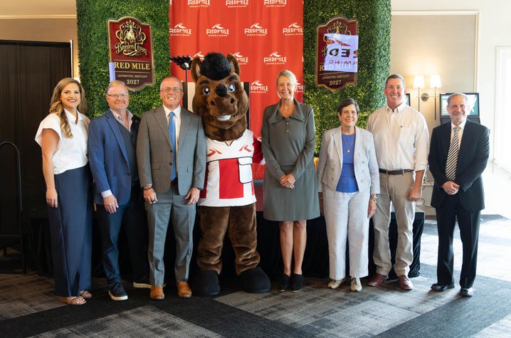 Seven people and a horse mascot stand together for a group photo in front of a red Red Mile Gaming & Racing backdrop and two “Breeders Crown Red Mile Lexington, Kentucky 2027” signs framed by green hedge panels. 