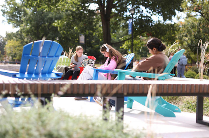 Three students sit in colorful Adirondack chairs outdoors, with two working on laptops beneath shade trees.