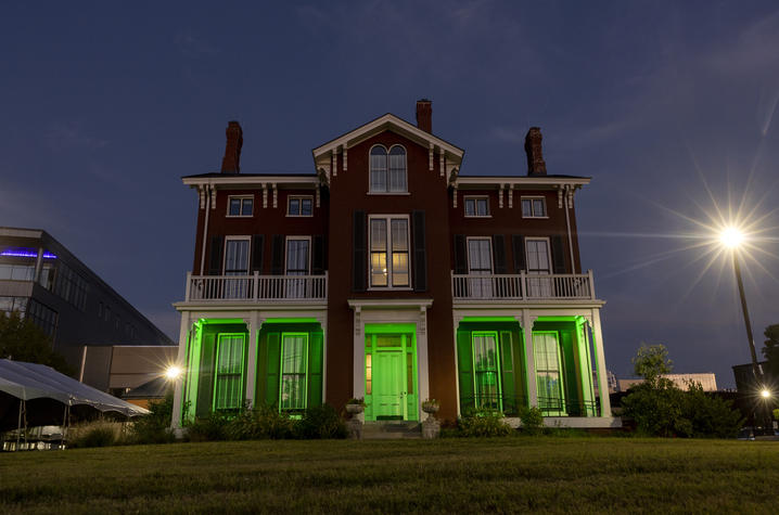 UK goes green Historic three-story red-brick house illuminated with green lighting along the front porch and entrance at dusk. The building features white columns, balconies, tall shuttered windows, and multiple chimneys. A grassy lawn stretches across the foreground.