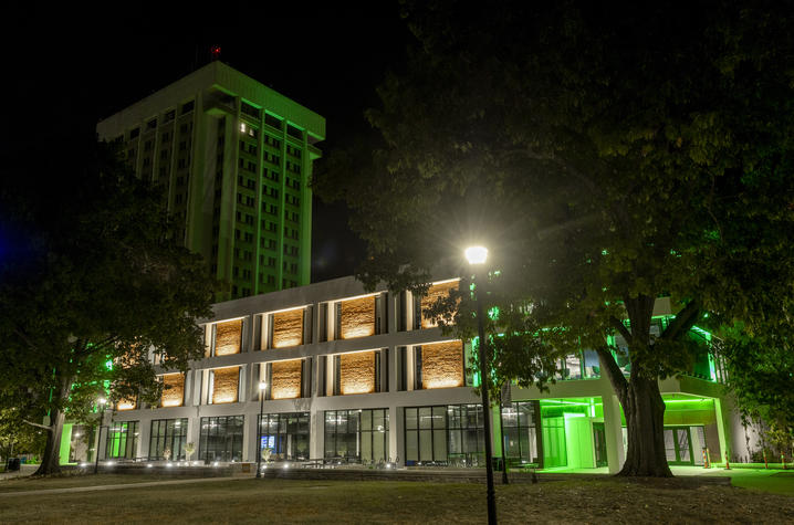 UK goes green Modern campus building illuminated with green accent lighting at night, with a taller residence hall glowing green in the background. Large trees frame the scene, while exterior lights highlight the building’s white facade, glass windows.