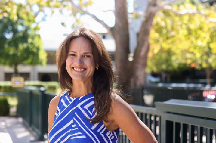 A person in a blue-and-white patterned top stands outdoors beside a railing, with trees and buildings in the background.