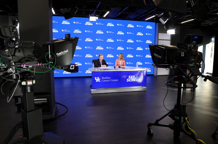 Photo of Mark Evers and Lee Anne Pope in studio. A television studio set with two people seated behind a blue-branded desk, viewed between two professional broadcast cameras in the foreground. Large teleprompter screens, studio lighting equipment, and cables surround the set.