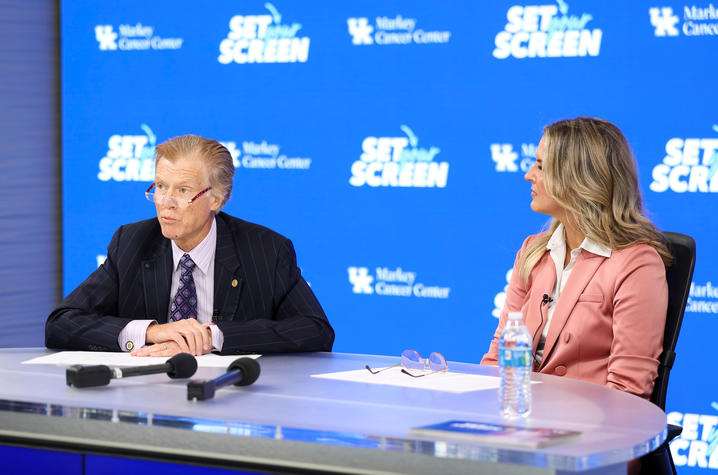 Photo of Mark Evers and Lee Anne Pope in studio. Two people sit at a curved desk equipped with microphones and papers in front of a blue backdrop featuring repeated University of Kentucky Markey Cancer Center logos and "Set Your Screen" campaign branding.