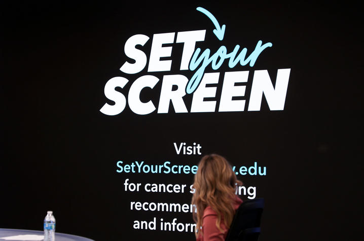 Photo of Lee Anne Pope and 'Set Your Screen' logo A large screen displays the “Set Your Screen” campaign logo above a website address and text encouraging visitors to find cancer screening recommendations and information. In the foreground, a person seated on a stage is viewed from behind a table.