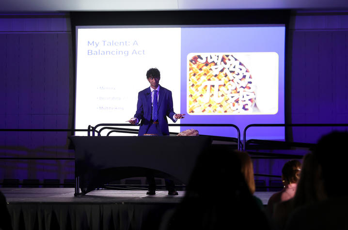 A speaker stands behind a table giving a presentation as a slide featuring colorful mahjong tiles is projected behind them.