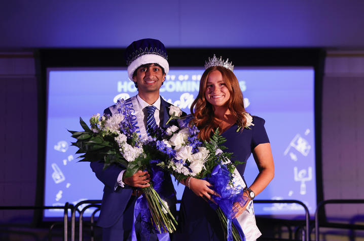 Two people wearing Homecoming crowns and sashes pose together while holding bouquets of flowers.