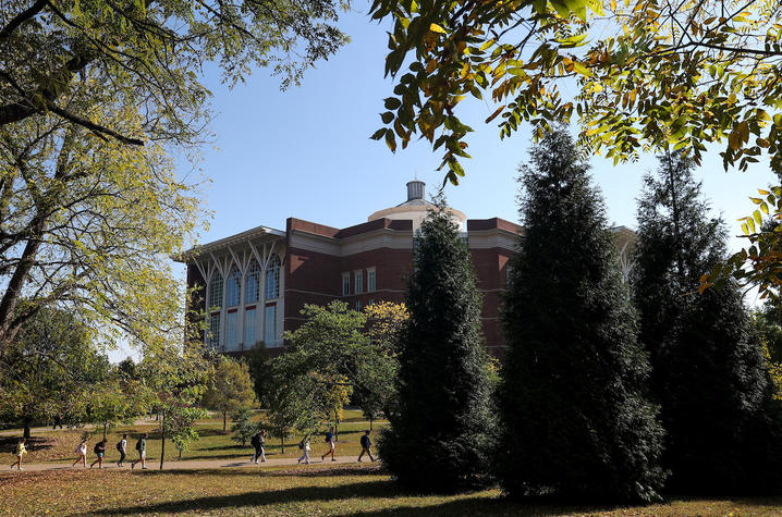 People walk along a tree-lined path outside a red brick campus building, framed by green and yellow autumn leaves under a clear blue sky.
