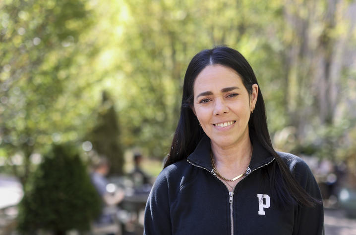 Person with long dark hair wearing a black zip-up jacket, standing outdoors in a landscaped setting.