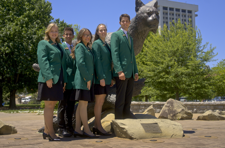 Five people standing lined up on the UK campus in front of a wildcat statue, wearing green 4-H formal jackets.