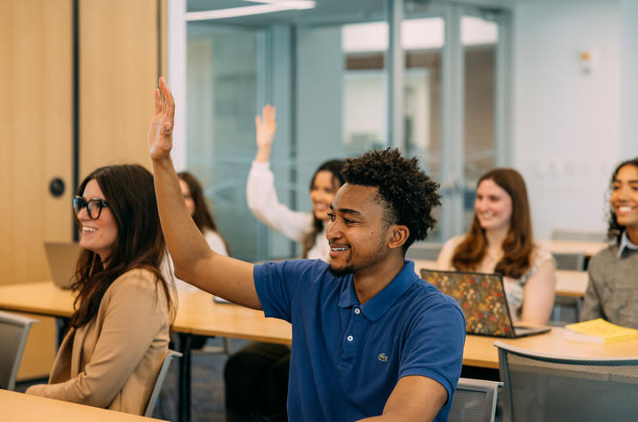 students sit at desks in a classroom and several have their hands raised