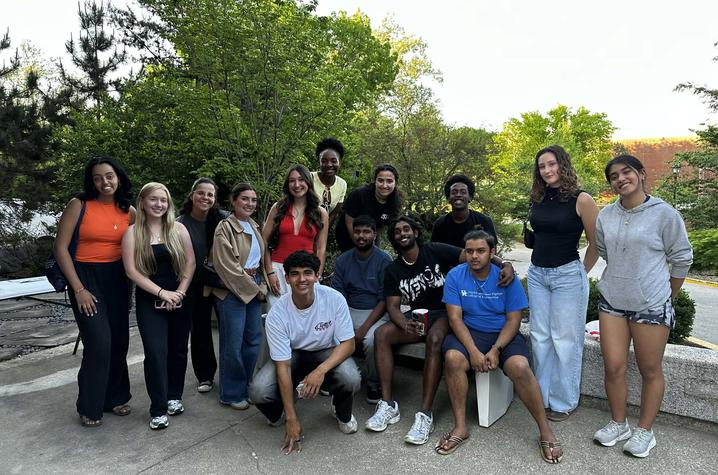 photo of ISLT A group of young adults pose outdoors, standing and sitting around a bench with leafy trees behind them.