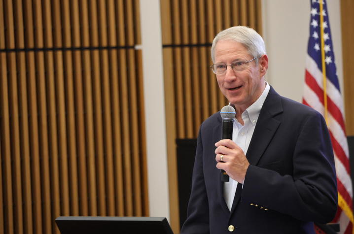 Person speaking into a handheld microphone at a podium in an indoor event space. An American flag is visible in the background, along with wood-paneled walls and presentation equipment.