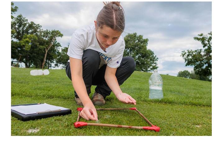 photo of student sampling turfgrass Person using a square sampling frame to examine grass in an outdoor field, with a clipboard and water container nearby.