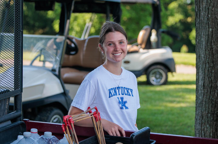 photo of UK graduate student Grace Ferry Person standing beside a utility vehicle in a grassy outdoor area, with field research equipment visible in the vehicle bed. The person is wearing a white T-shirt with the text “KENTUCKY UK,” and golf carts and trees can be seen in the background.