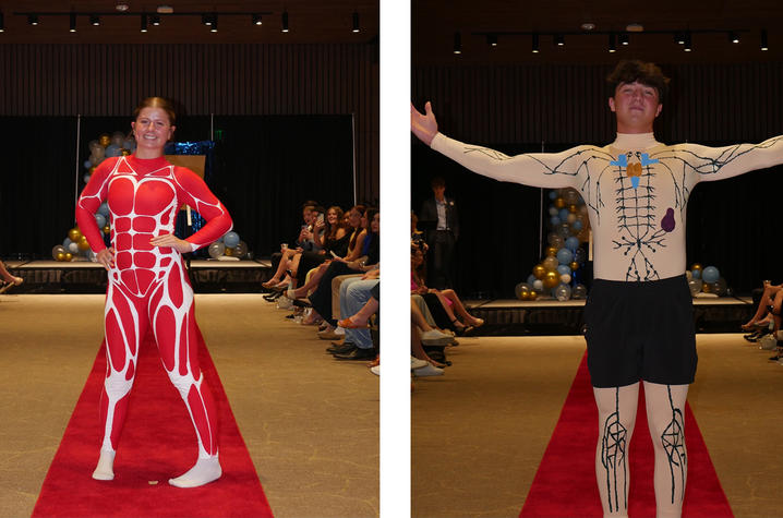 Two people walk a red carpet runway, each wearing clothing printed with anatomical designs — one in a red outfit showing muscle patterns and one in a light‑colored outfit with bone and organ illustrations.