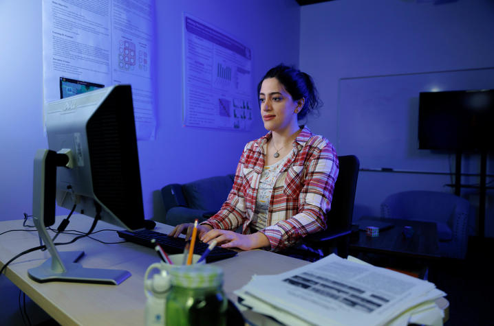  A person seated at a desk works on a computer in a dimly lit office with blue lighting. Posters or charts hang on the wall behind them, and papers and a drink sit on the desk in the foreground.