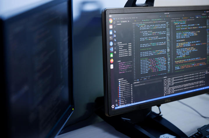  A close-up view of two computer monitors in a dimly lit workspace; one monitor displays multiple windows of code with syntax highlighting, while the other is blurred and mostly dark.