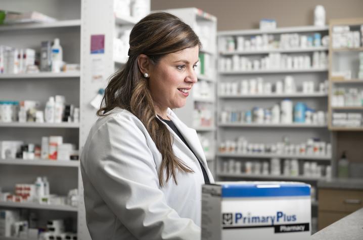 Pharmacist in a white lab coat standing behind a pharmacy counter with shelves of medication in the background.