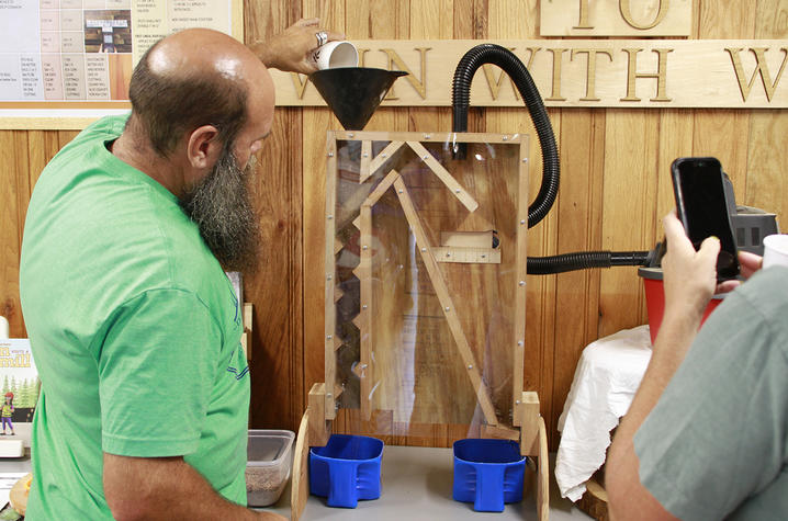 A person pours material from a cup into a funnel atop a transparent, wood-framed seed-sorting machine during a workshop demonstration.