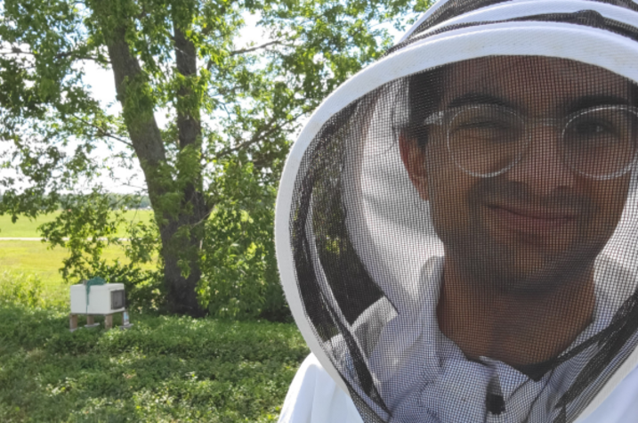 Student conducts research outdoors as part of University of Kentucky CURE fellowship program Person wearing a beekeeping suit with a mesh hood, standing outdoors near a tree and a beehive in a grassy area.