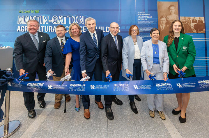 Group of university leaders, faculty and supporters stand behind a ribbon at a ceremonial opening event inside a campus building with blue signage in the background.