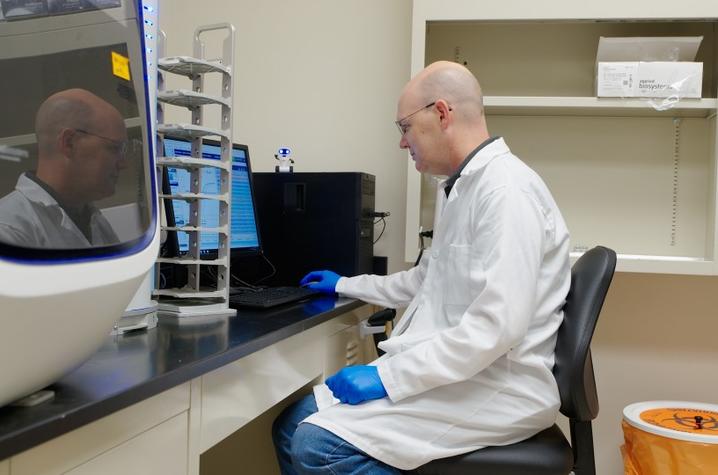 A person in a lab coat and gloves sits at a computer workstation in a laboratory.