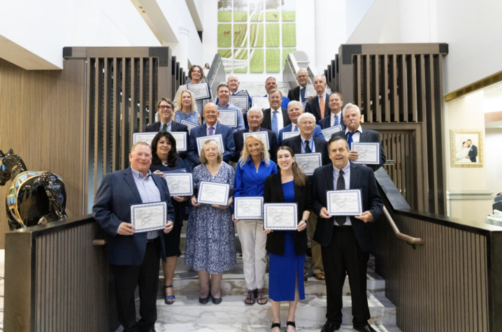 Alumni award recipients stand on a staircase holding certificates during a recognition ceremony in a building lobby.