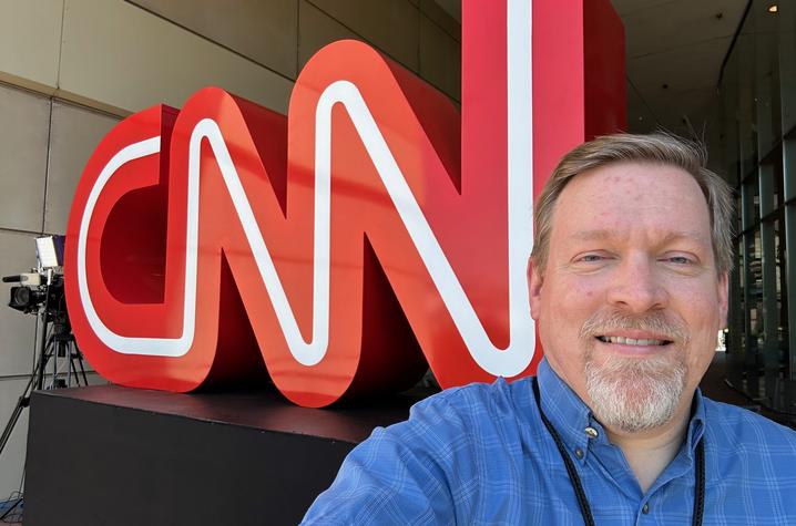 A man in a blue button-down shirt smiles for a selfie in front of a large red CNN logo outside the CNN Center.