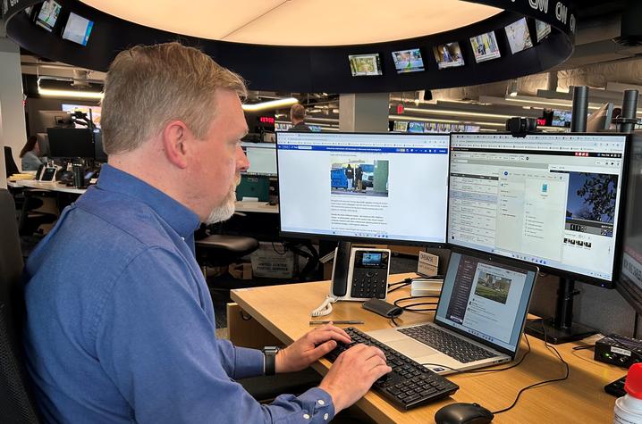 A man in a blue button-down shirt works at a desk in the CNN newsroom, typing on a keyboard while viewing news content across multiple computer monitors. Television screens line a circular display overhead, and a laptop and office phone on the desk.
