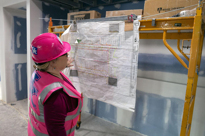 A person wearing a bright pink safety vest and a pink hard hat stands inside an unfinished construction area, looking at a large building plan or blueprint taped to a scaffold.