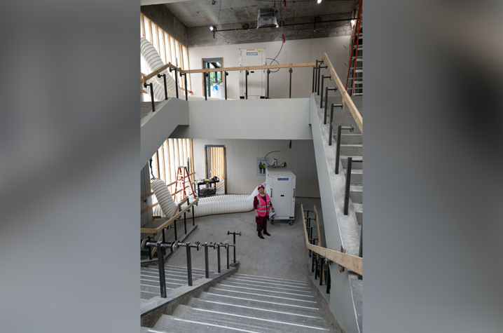 An unfinished multilevel stairwell with concrete steps, metal railings and construction equipment. A person in safety gear stands on the lower level.