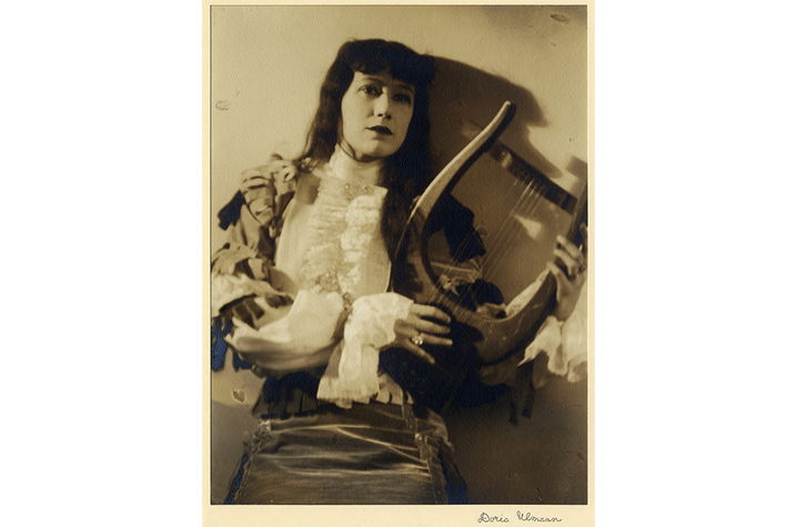 image of Angna Enters by Doris Ulmann Sepia-toned portrait of a person seated in a theatrical costume, holding a lyre. The subject faces forward, framed by patterned fabric and expressive textures, with a faint signature or caption visible at the bottom of the image.
