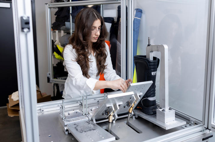 Working at ArcWear, a division of Kinectrics Individual wearing clear safety glasses adjusts a testing setup inside a large enclosed lab apparatus. A black work boot is mounted on a metal fixture to the right, while angled metal plates and small debris sit on the testing surface.