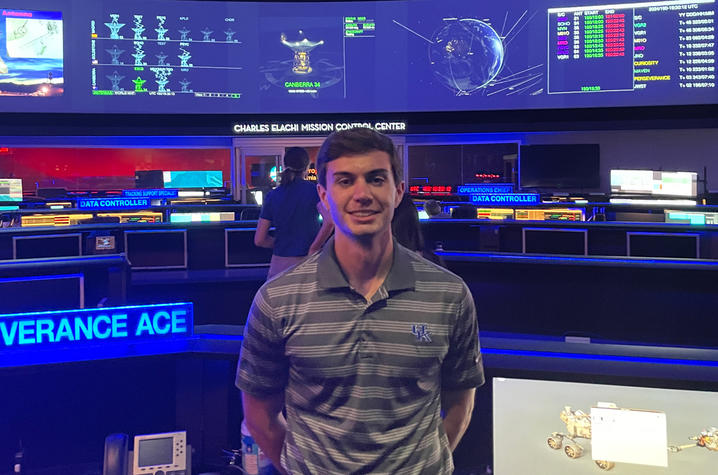 White male in dark grey and light grey striped collared shirt, standing in a NASA mission control room