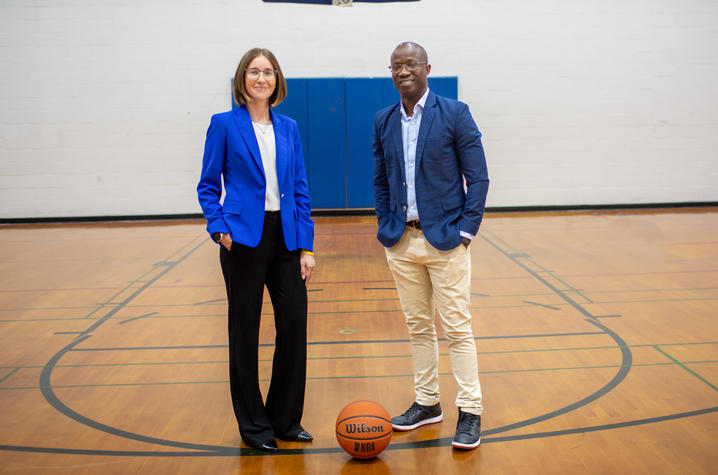 Photo of Amanda Duncan and Tshepang Tshube Two people stand on a gym basketball court beneath a hoop, with a basketball on the floor between them.