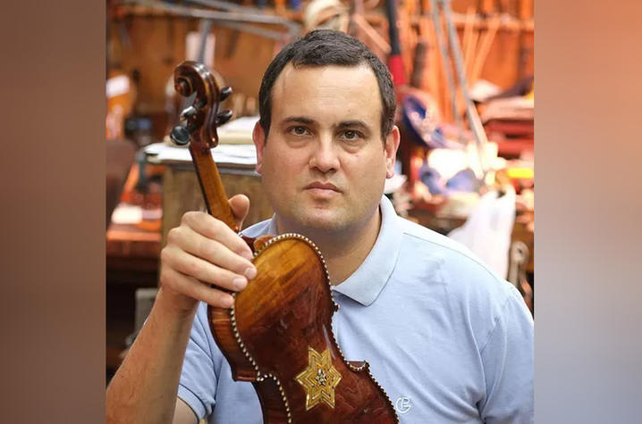A person wearing a light blue shirt holds up a violin in a workshop. The back of the violin features an intricate inlaid six‑pointed star design. Various tools and instruments are visible in the background.