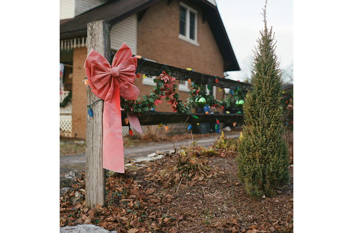 A large red bow is tied around a wooden post in a yard with winter foliage, with a house in the background decorated with colorful holiday lights and greenery.