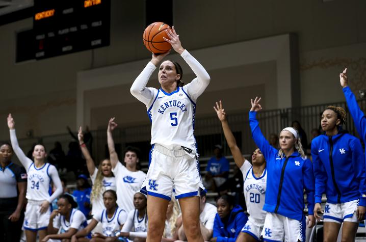 blair green in a kentucky basketball uniform shooting a three. her teammates are standing behind her cheering her on.