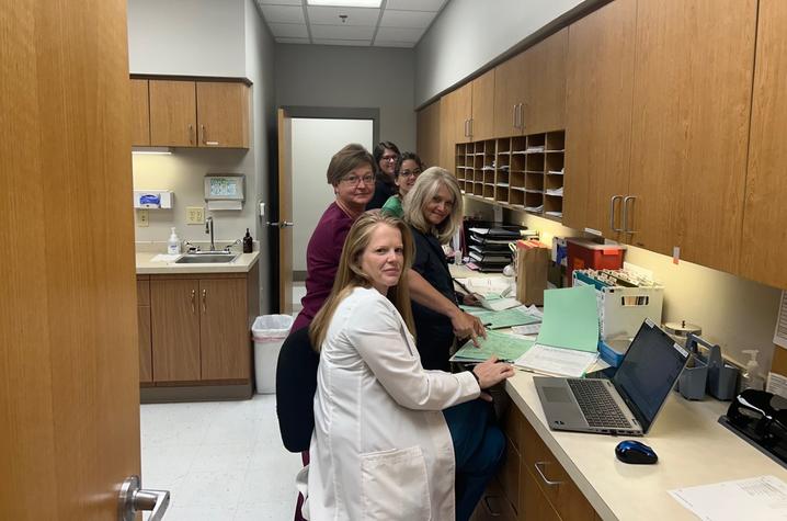 photo of outreach team in bourbon county Five people gather at a workstation in a medical office, surrounded by wooden cabinets, paperwork, patient files and a laptop; one person in a white coat sits at the counter while the others stand nearby.