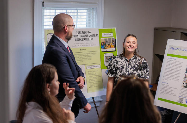 photo of Macy French A presenter stands beside research posters and speaks to a small group gathered in a room during a poster presentation session.